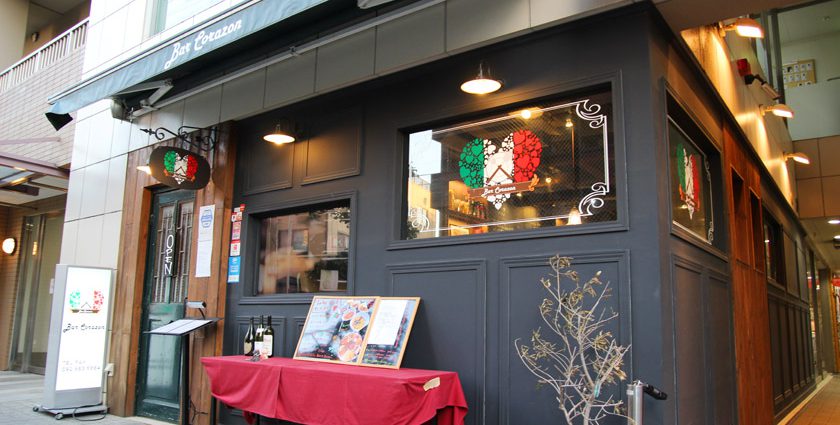 唐人町商店街にあるイタリア＆ラテン料理レストラン、バル・コラソンの外観です。店舗の壁は黒色で入り口のドア枠は木製です。窓ガラスにイタリア国旗の配色のハートマークの絵が描かれています。入り口付近には赤いテーブルクロスがかけられた長机があり、ワインボトル、メニュー板が置かれています。