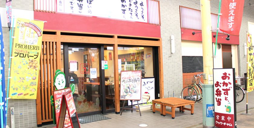 唐人町商店街にあるお惣菜、お弁当のお店クックチャムです。「おかずとお弁当」と書かれたサインが立っています。入り口はガラスのドアで木目調の枠があります。