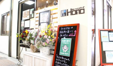 唐人町商店街にあるカレー＆フレンチトースト Honoの外観です。白色の壁に「カレー＆ハンバーグ　HONO」と書いてあります。いくつかの花束が棚の上に置かれています。「テイクアウトは厨房側の扉にて承ります」と書かれた看板が立てかけられています。