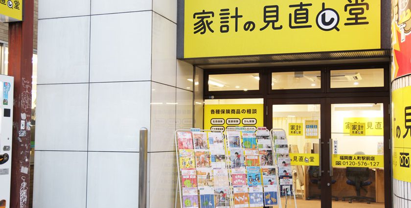 唐人町商店街にある家計の見直し堂 福岡唐人町駅前店の外観です。壁に黄色い看板に家計の見直し堂と書かれています。入り口にはパンフレットラックがあり、たくさんのチラシが配置されています。